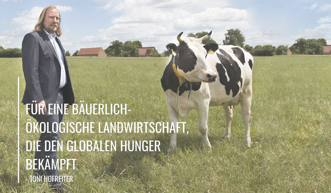 Toni steht neben einer Kuh. Auf dem Bild steht: "Für eine bäuerlich-ökologische Landwirtschaft, die den globalen Hunger bekämpft." Im Hintergrund stehen Bäume und es ist eine Wiese zu sehen.