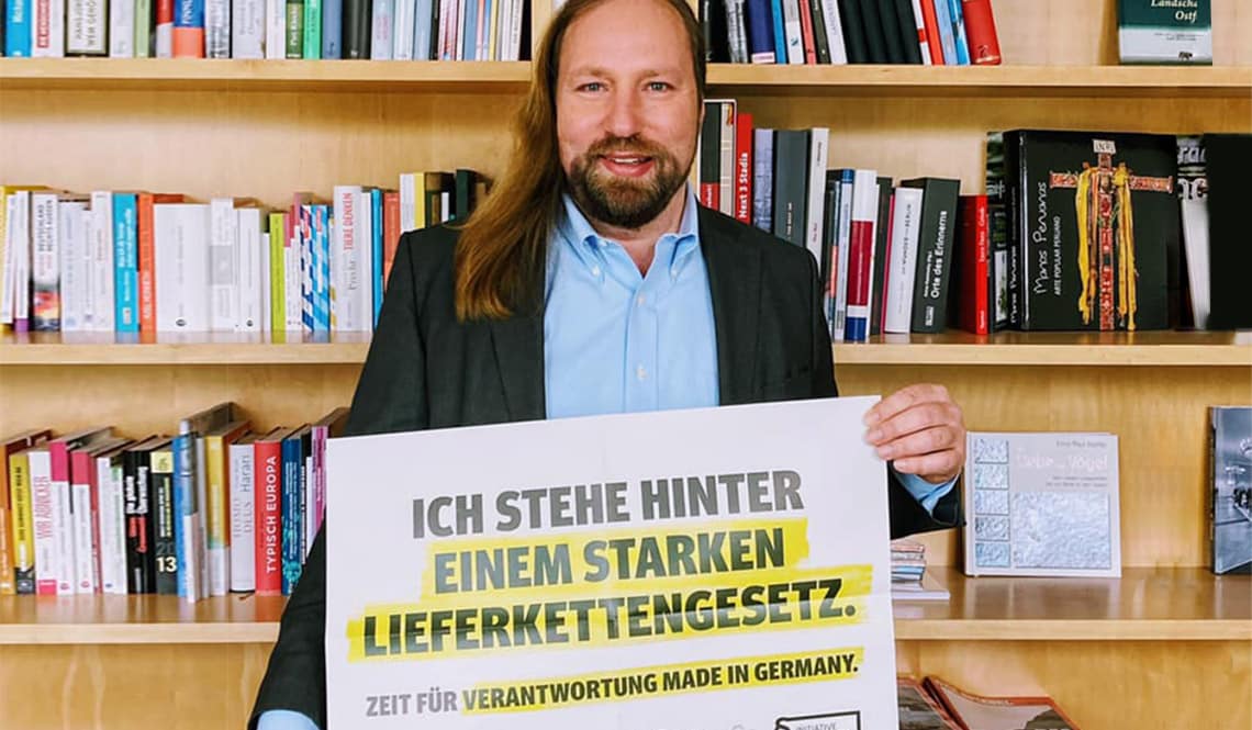 Toni Hofreiter steht vor einem Bücherregal. Er hält ein Schild in der Hand auf dem Schild steht: "Ich stehe hinter einem starken Lieferkettengesetz".